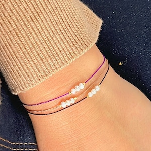 May include: Three delicate bracelets with small white pearls and gold beads on a brown, black, and purple string. The bracelets are worn on a person's wrist.