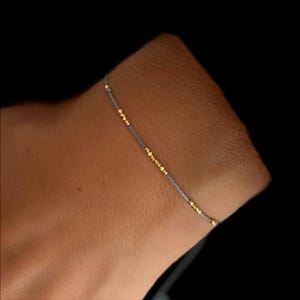 Op de afbeelding: Een fijne armband met kleine gouden kralen geregen op een dun, grijs koord. De armband heeft een minimalistisch ontwerp, ideaal voor dagelijks gebruik. De gouden kralen zijn gelijkmatig verdeeld.