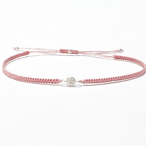 Könnte beinhalten: Ein zartes Armband aus geflochtenem rosa Band mit einem kleinen silbernen Anhänger mit einem klaren Kristall.
