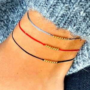 Könnte beinhalten: Drei übereinanderliegende Armbänder mit goldenen Perlen. Das obere Armband ist grau, das mittlere Armband ist rot und das untere Armband ist schwarz.