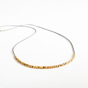 Può includere: Delicata collana con una fila di piccole perline dorate sfaccettate. Le perline sono infilate su un sottile cordoncino grigio. Una piccola chiusura dorata a forma di stella assicura la collana. Su sfondo bianco.