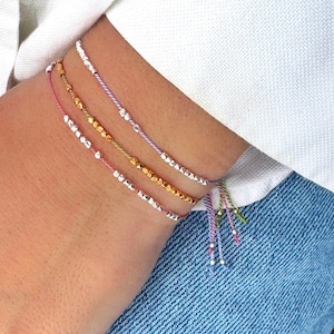 Könnte beinhalten: Vier übereinanderliegende Armbänder mit silbernen und goldenen Perlen auf bunter Schnur. Die Armbänder sind mit einem Knoten gebunden und haben lange Schnüre, die vom Knoten hängen.