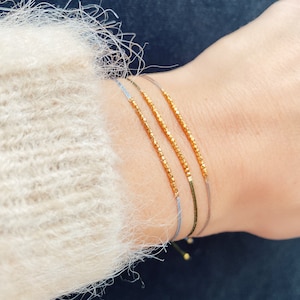 Könnte beinhalten: Drei goldene Perlenarmbänder an einem Handgelenk. Die Armbänder bestehen aus kleinen, goldenen, facettierten Perlen und sind auf dünnen, grauen Schnüren aufgefädelt.