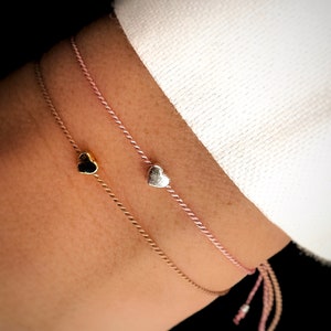 May include: Two delicate bracelets with heart charms. One bracelet is pink and the other is brown. Both bracelets have a heart charm at the end.