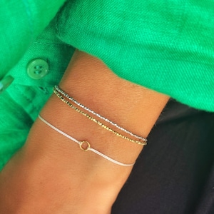 May include: Three bracelets on a wrist. The top bracelet is silver and gold, the middle bracelet is gold, and the bottom bracelet is white with a gold ring charm.