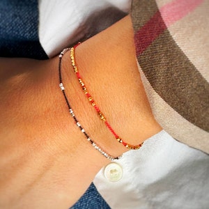 May include: Three bracelets stacked on a person's wrist. The top bracelet is red string with gold beads. The middle bracelet is gold beads. The bottom bracelet is black string with silver beads.