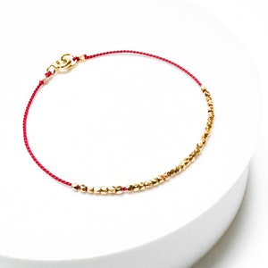 May include: A red string bracelet with gold beads. The beads are small and have a faceted design.