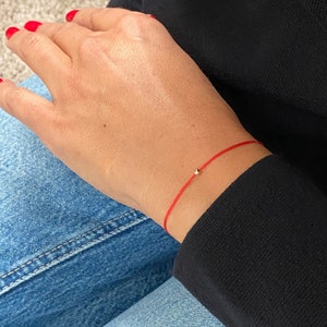 Könnte beinhalten: Ein rotes Armband mit einer kleinen goldenen Perle. Das Armband wird am Handgelenk getragen.
