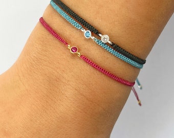 May include: Three bracelets with different colored cords and small gemstone accents. The top bracelet has a black cord with a blue and white gemstone. The middle bracelet has a blue cord with a white gemstone. The bottom bracelet has a red cord with a red gemstone.