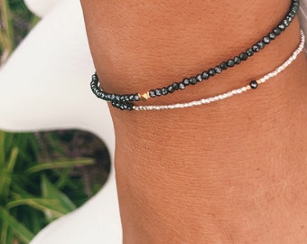 Puede incluir: Dos pulseras en la muñeca de una persona. La pulsera superior está hecha de cuentas negras con detalles dorados. La pulsera inferior está hecha de cuentas plateadas con detalles dorados.