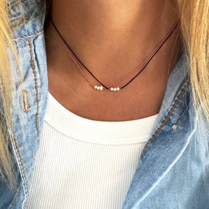 Puede incluir: Un collar delicado con dos hebras de cordón, una negra y otra rosa, con pequeñas perlas blancas espaciadas a lo largo de las hebras.