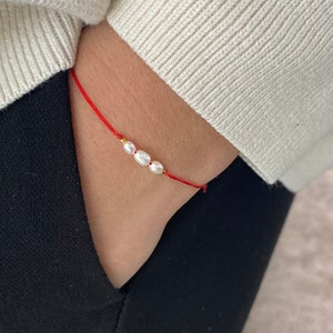 May include: A delicate bracelet featuring three white pearls and gold accents strung on a thin red cord. The bracelet is worn on a wrist, with a cream-coloured sweater and black trousers in the background.