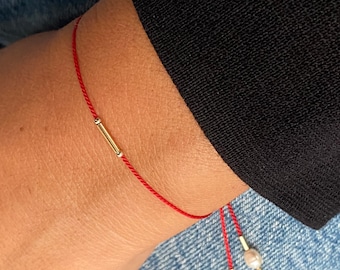 May include: A red string bracelet with a gold bar charm and two small pearl charms. The bracelet is tied with a knot and the ends are left long.