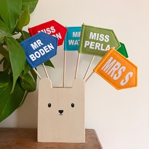 Puede incluir: Un portalápices de madera con forma de oso con varias banderas de fieltro de colores. Las banderas, en rojo, azul, verde y naranja, tienen texto blanco con nombres como "Miss Perla" y "Mr Boden".