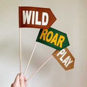 Op de afbeelding: Drie vlaggetjes van vilt met houten stokjes. De vlaggetjes zijn bruin, groen en bruin. De tekst op de vlaggetjes is "WILD", "ROAR" en "PLAY".