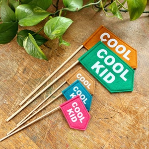 Può includere: Quattro bandiere di feltro con bastoncini di legno. Le bandiere sono di colori diversi e hanno il testo "Cool Kid" scritto su di esse. Le bandiere sono su una superficie di legno.