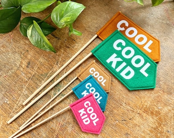 Może przedstawiać: Cztery flagi z filcu z drewnianymi patyczkami. Flagi są w różnych kolorach i mają napisane na nich "Cool Kid". Flagi leżą na drewnianej powierzchni.