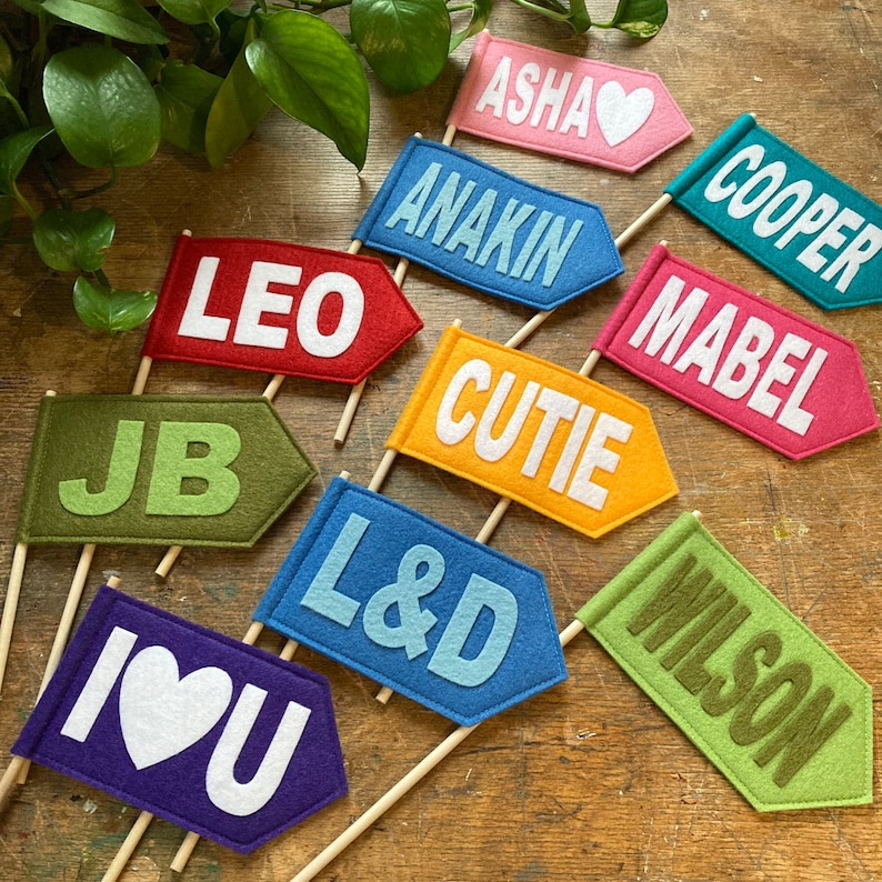 Op de afbeelding: Tien kleurrijke viltvlaggen met verschillende namen in het wit geschreven. De namen zijn: Asha, Anakin, Leo, JB, L&D, Cutie, Mabel, Wilson, Cooper en Ik hou van je.