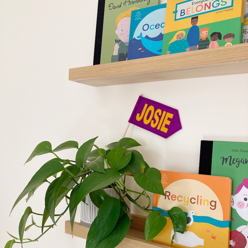 Op de afbeelding: Een paarse vlag van vilt met de naam "JOSIE" erop geschreven in gele letters. De vlag hangt aan een witte muur met een houten plank erboven. Er staan boeken op de plank en er staat een plant in een pot onder de vlag.