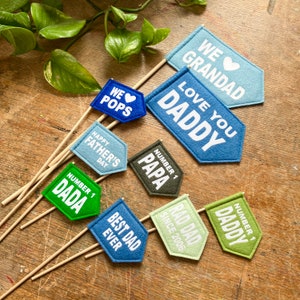 Peut inclure: Une collection de fanions en feutrine sur des bâtons en bois de différentes couleurs, dont bleu, vert et olive. Les fanions portent des inscriptions blanches : WE LOVE GRANDAD, LOVE YOU DADDY, WE LOVE POPS, HAPPY FATHER'S DAY, NUMBER 1 PAPA, NUMBER 1 DADA, BEST DAD EVER, RAD DAD SINCE 2006 et NUMBER 1 DADDY.