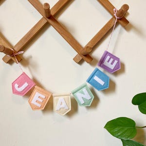 Puede incluir: Una guirnalda de fieltro que deletrea el nombre "JEANIE" en letras mayúsculas blancas. Cada letra está centrada en un banderín de fieltro pentagonal de colores pastel, incluyendo rosa, naranja, crema, verde menta, azul y morado, unidos por un cordel.