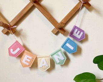 Könnte beinhalten: Ein Filzbanner, das den Namen "JEANIE" in weißen Großbuchstaben buchstabiert. Jeder Buchstabe ist auf einem fünfeckigen Filzwimpel in Pastellfarben wie Rosa, Orange, Creme, Mintgrün, Blau und Lila zentriert und an einer dünnen, rosa-weißen Schnur befestigt.