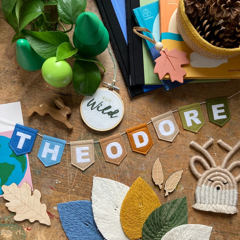 Puede incluir: Un bander&iacute;n de fieltro con el nombre Theodore en letras blancas sobre banderines coloridos. El bander&iacute;n est&aacute; rodeado de otros elementos decorativos, incluyendo un conejo de madera, un peque&ntilde;o aro con las palabras "Run Wild" bordadas en &eacute;l, y una variedad de hojas y otros elementos naturales.