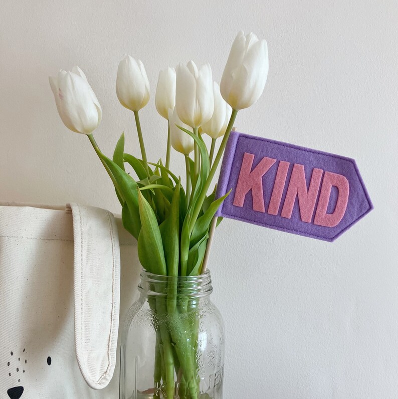 Op de afbeelding: Een boeket witte tulpen in een glazen vaas met een paarse viltvlag waarop "KIND" in roze letters staat.