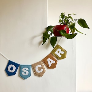 Puede incluir: Un banderín de fieltro con la palabra "OSCAR" en letras blancas sobre banderines coloridos. Los banderines son en tonos azules, beige y verdes, ensartados con una cuerda blanca. Una maceta roja con una planta verde en el fondo.