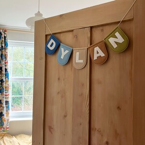 Puede incluir: Una pancarta de fieltro con el nombre "DYLAN" escrito en letras blancas sobre banderines de fieltro de colores. La pancarta est&aacute; colgada en una puerta de madera.