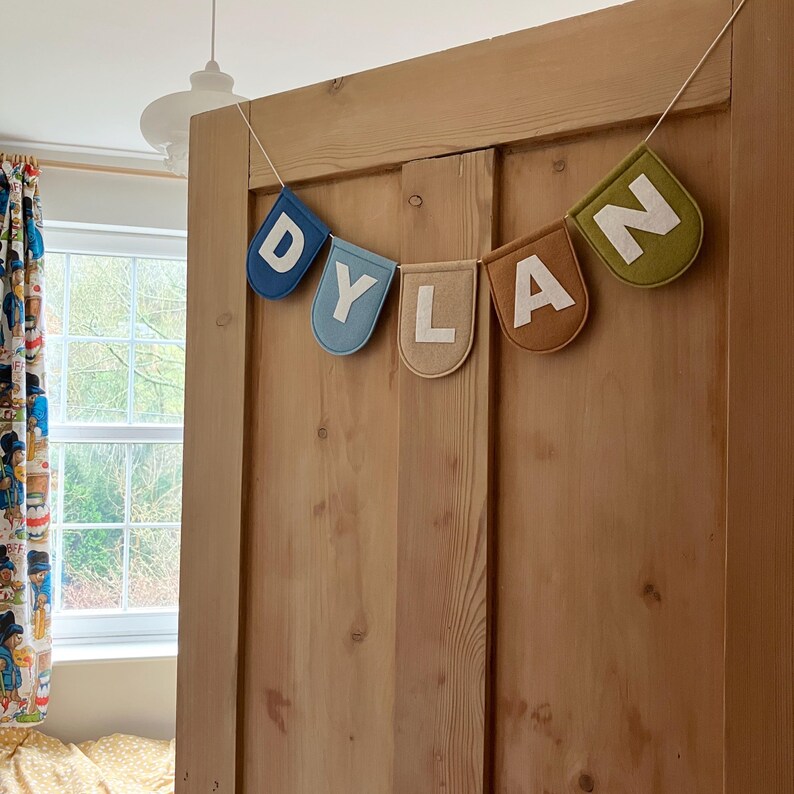 Puede incluir: Una pancarta de fieltro con el nombre "DYLAN" escrito en letras blancas sobre banderines de fieltro de colores. La pancarta est&aacute; colgada en una puerta de madera.