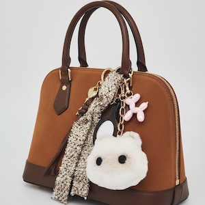 Może przedstawiać: Brązowa torebka o zaokrąglonym kształcie, z górnym uchwytem i zapięciem na zamek błyskawiczny. Torebka ozdobiona jest brelokiem z białym, puszystym misiem, różowym balonowym psem i wzorzystym szalikiem.