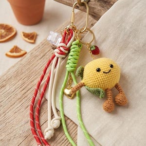 May include: A handmade keychain featuring a yellow crocheted lemon with a smiling face, green leaf, and brown legs. The keychain includes red, white, and green cords, a gold-colored clasp, and a small strawberry charm. The background includes a wooden surface and a small potted plant.