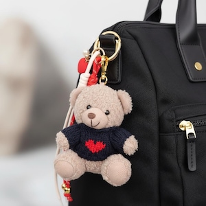 May include: A beige teddy bear keychain with a navy blue sweater featuring a red heart design. The plush toy is attached to a gold-colored keyring with a red and white decorative cord, hanging from a black bag.