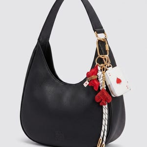 May include: Black leather hobo bag with a curved shape. The bag features a gold-tone chain and a charm with playing card motifs, including a red heart and a white card with a red heart. A red and white rope tassel hangs from the charm.