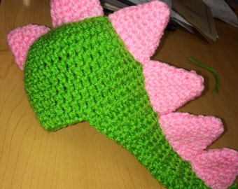 May include: A green and pink crocheted dinosaur tail with a long, curved body and a series of pink spikes.