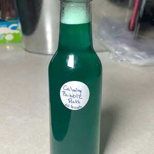 May include: A clear glass bottle filled with green liquid and topped with a black cap. The bottle has a white label with the words "Calming Bubble Bath Kid Friendly" written in blue ink. The liquid has a foamy top.
