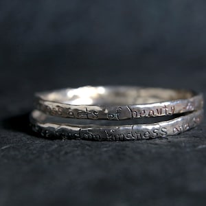 May include: Two silver rings stacked on top of each other. The top ring has the inscription "small acts of beauty" and the bottom ring has the inscription "everyday kindness and love".