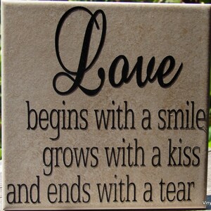 May include: A beige ceramic tile with black lettering that reads "Love begins with a smile grows with a kiss and ends with a tear".