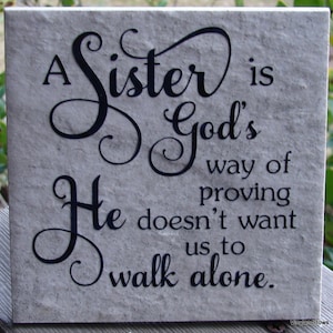 May include: A decorative stone tile with a black cursive quote that reads "A Sister is God's way of proving He doesn't want us to walk alone."