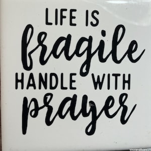 May include: White square tile with black text that reads "LIFE IS FRAGILE HANDLE WITH PRAYER". The text is in a handwritten style. The tile has a glossy finish.
