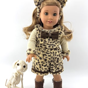 May include: A doll with long brown hair wearing a leopard print headband, a matching leopard print romper with a brown bow, and brown boots. Beside the doll stands a small white plastic dog skeleton with black eyes and a detailed rib cage structure.