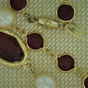 May include: Gold-toned necklace featuring alternating faux pearls and faceted, deep red gemstones. The necklace has a chain link design with a magnetic clasp and a small tag with text.
