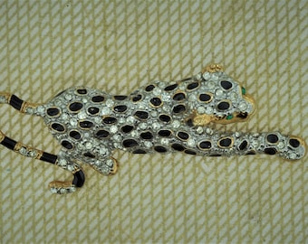 May include: A jeweled leopard brooch. The brooch features a gold-toned metal body with black enamel spots and stripes. The leopard's body is covered in clear rhinestones, with green gem eyes.