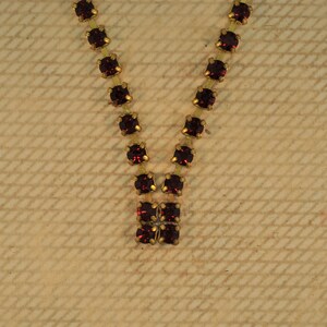 May include: A gold-toned necklace with a V-shaped design. The necklace features square, deep red gemstones set in gold-colored metal. The gemstones are evenly spaced along the chain, creating a symmetrical pattern.