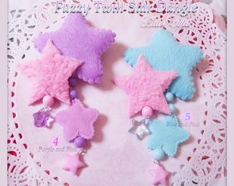 May include: Two fuzzy star hair clips with dangling charms. The stars are pink and blue, with purple and pink charms on one clip and blue and pink charms on the other. The text "Fuzzy Twin Star Dangle Hair Clips" is at the top of the image.