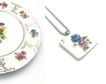 May include: A white porcelain plate and pendant necklace. The plate features a floral bouquet in the center with gold filigree around the rim. The pendant has a floral design with the number "5" and hangs on a silver ball chain.