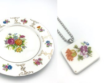 May include: A white porcelain plate with a gold trim and floral design. The plate features a bouquet of colorful flowers in the center and smaller floral motifs around the edge.  A white porcelain pendant necklace with a square floral design.