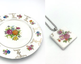 May include: A white ceramic plate with a gold rim and floral design. The plate features a central bouquet of pink, orange, yellow, and blue flowers. The plate is surrounded by a repeating pattern of smaller floral motifs. The image also shows a square white ceramic pendant with a floral design on a silver chain.