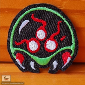 May include: Black and green embroidered patch of a video game character with red eyes and white pupils. The character has a green rim around its head and red teeth.